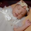 Close-up of a sleeping South Asian baby dressed in a white gown and headband, exuding innocence and peace.