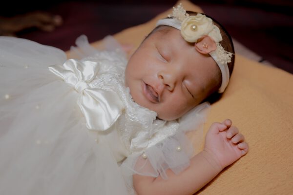 Close-up of a sleeping South Asian baby dressed in a white gown and headband, exuding innocence and peace.